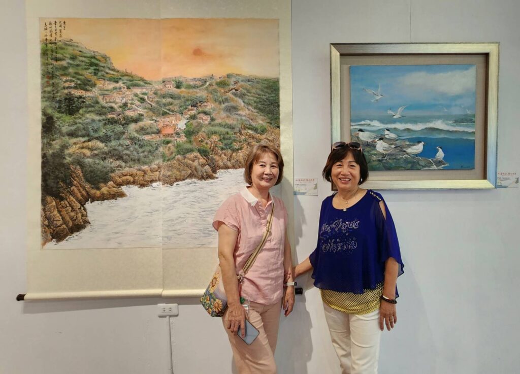 ▲馬祖參展藝術家劉玉燕(左)與曹賽娥,在各自的彩墨與油畫作品前開心為來賓導覽,馬祖的山海風情一覽無遺。(圖/大媒體新聞網 攝)