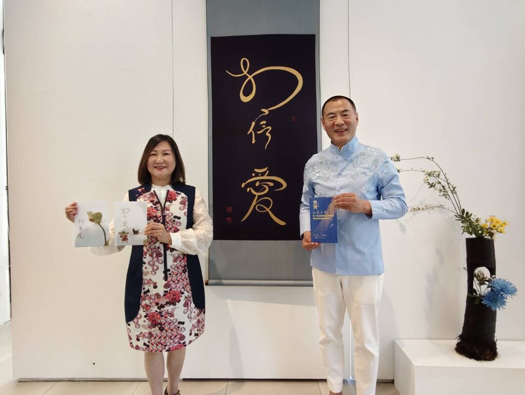 ▲大愛光老師周瑞宏（右）及師娘黃玉華分持展覽手冊及文創品（書法孫端吉、繪畫蔡悅璐），在孫端吉教授作品「相信愛」前合影。（記者 辛澎祥/攝）