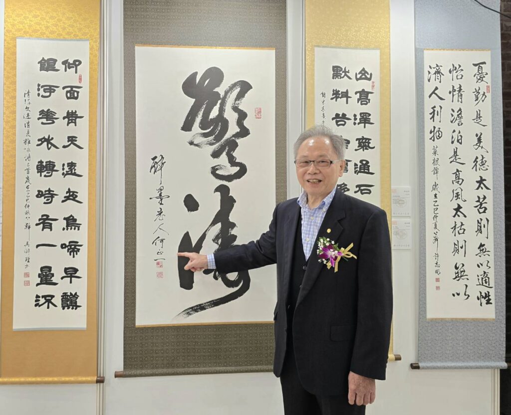 ▲淨心書會創會會長何正一在其展出作品前與作品合影，呈現其特有的書藝風格。（記者 詹佳綺/攝）