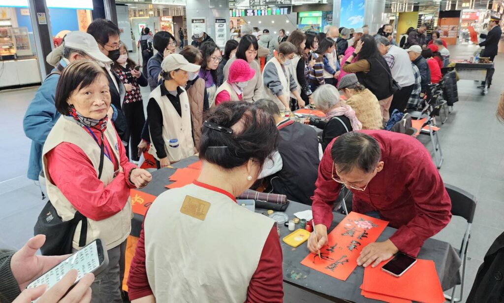 ▲民眾及旅客在臺鐡西側廣場踴躍向書畫老師們排隊索取春聯的熱鬧景況，讓行之多年的協會與臺鐡合辦寫春聯活動再現高峰。（圖/大媒體新聞網 提供）
