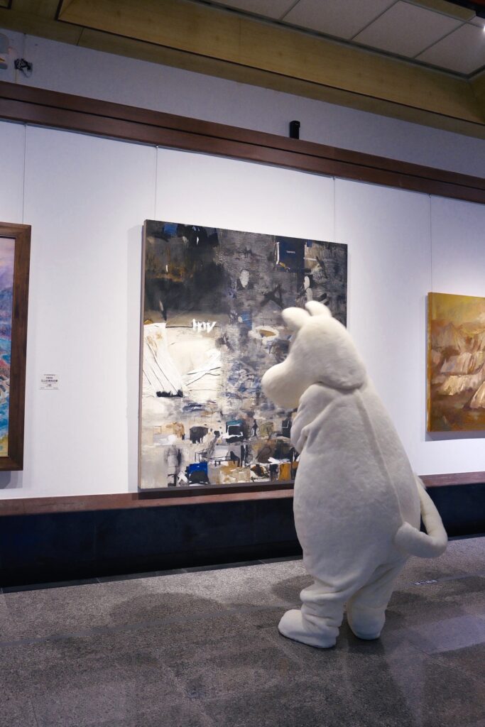 ▲姆明人偶來到港區藝術中心，駐足在全國百號油畫大展的畫作前。（圖/文化局提供）